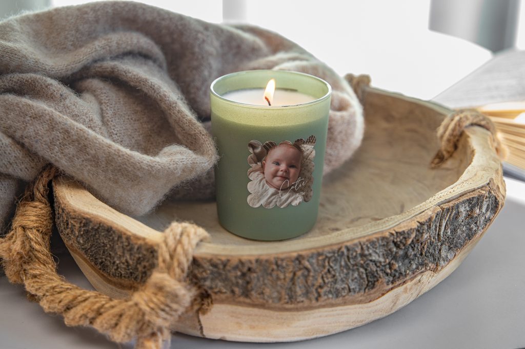 Grüne Duftkerze mit Kinderfoto auf dem Etikett auf einem Holztablett neben einer Wolldecke
