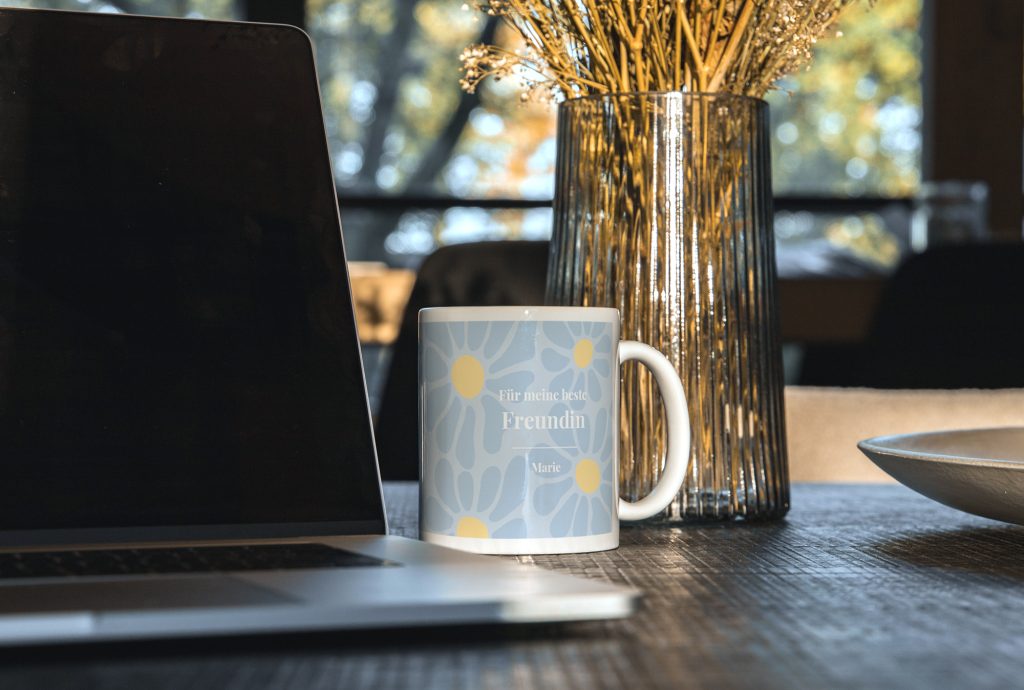 Hellblaue Tasse mit Blumenmuster und Namen neben einem Laptop auf einem Holztisch