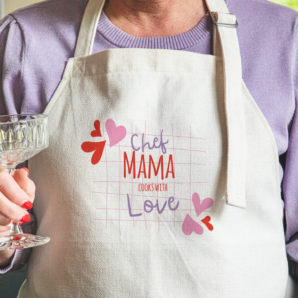 Frau trägt eine helle Kochschürze mit Herzchen und dem Aufdruck „Chef Mama“